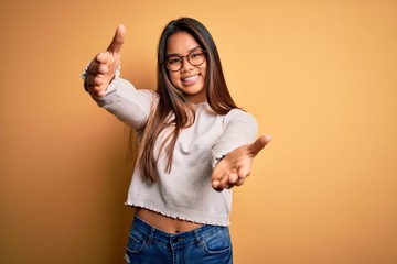 Young beautiful asian girl wearing casual sweater and glasses over yellow background looking at the camera smiling with open arms for hug. Cheerful expression embracing happiness.