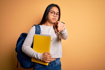 Young smart asian student girl wearing backpack holding notebook over yellow background pointing with finger to the camera and to you, hand sign, positive and confident gesture from the front