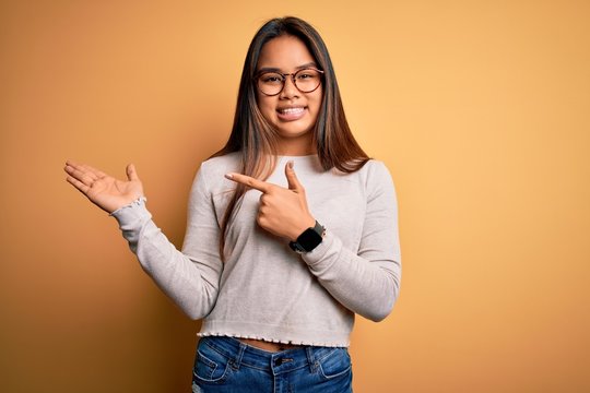 Young beautiful asian girl wearing casual sweater and glasses over yellow background amazed and smiling to the camera while presenting with hand and pointing with finger.