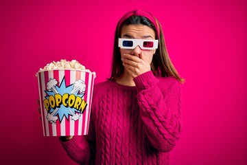 Young beautiful girl watching movie using 3d glasses eating box with popcorns shocked covering mouth with hands for mistake. Secret concept.