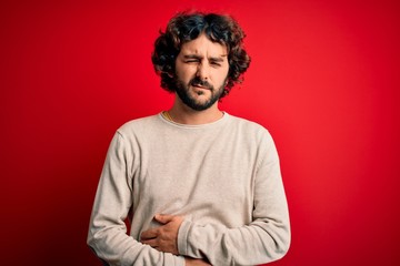 Fototapeta na wymiar Young handsome man with beard wearing casual sweater standing over red background with hand on stomach because indigestion, painful illness feeling unwell. Ache concept.