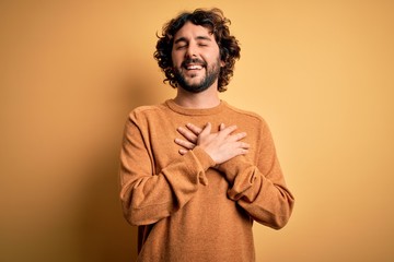 Young handsome man with beard wearing casual sweater standing over yellow background smiling with hands on chest with closed eyes and grateful gesture on face. Health concept.