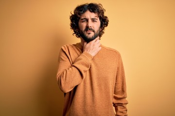 Young handsome man with beard wearing casual sweater standing over yellow background Touching painful neck, sore throat for flu, clod and infection