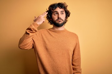 Young handsome man with beard wearing casual sweater standing over yellow background Shooting and killing oneself pointing hand and fingers to head like gun, suicide gesture.