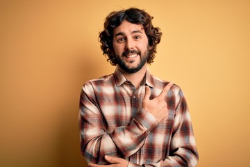 Young handsome man with beard wearing casual shirt standing over yellow background cheerful with a smile of face pointing with hand and finger up to the side with happy and natural expression on face