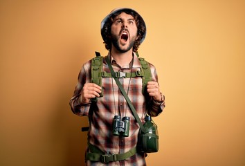 Young hiker man with curly hair and beard hiking wearing backpack and water canteen angry and mad screaming frustrated and furious, shouting with anger. Rage and aggressive concept.