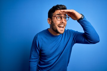 Young handsome man with beard wearing casual sweater and glasses over blue background very happy and smiling looking far away with hand over head. Searching concept.