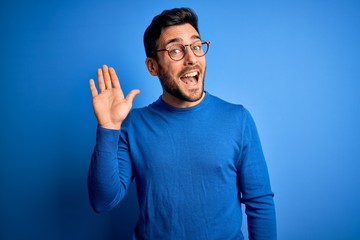 Young handsome man with beard wearing casual sweater and glasses over blue background Waiving...