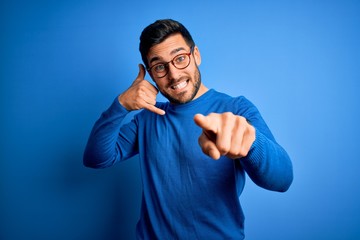 Young handsome man with beard wearing casual sweater and glasses over blue background smiling doing talking on the telephone gesture and pointing to you. Call me.