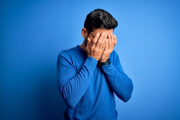 Young handsome man with beard wearing casual sweater and glasses over blue background with sad expression covering face with hands while crying. Depression concept.
