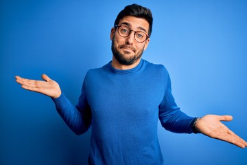 Young handsome man with beard wearing casual sweater and glasses over blue background clueless and confused expression with arms and hands raised. Doubt concept.