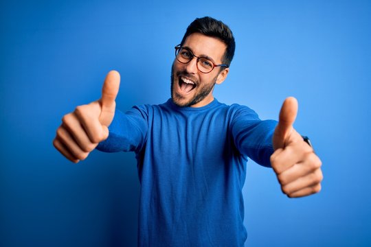 Young handsome man with beard wearing casual sweater and glasses over blue background approving doing positive gesture with hand, thumbs up smiling and happy for success. Winner gesture.