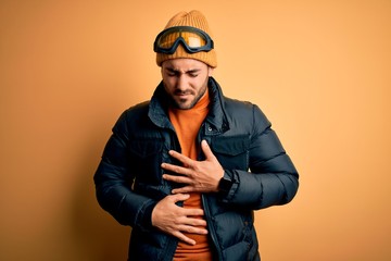 Young handsome skier man with beard wearing snow sportswear and ski goggles with hand on stomach because indigestion, painful illness feeling unwell. Ache concept.