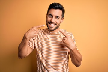 Young handsome man with beard wearing casual striped t-shirt over yellow background smiling...