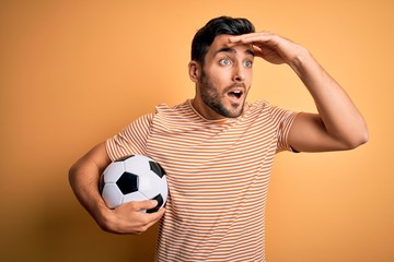 Handsome player man with beard playing soccer holding footballl ball over yellow background very...