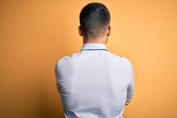 Young handsome businessman wearing tie and glasses standing over yellow background standing backwards looking away with crossed arms