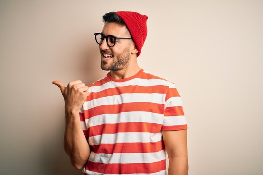 Young handsome man wearing casual striped t-shirt and glasses over white background smiling with happy face looking and pointing to the side with thumb up.
