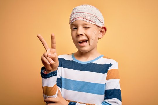 Young little caucasian kid injured wearing medical bandage on head over yellow background smiling with happy face winking at the camera doing victory sign. Number two.