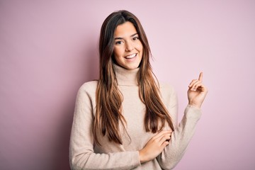Young beautiful girl wearing casual turtleneck sweater standing over isolated pink background with a big smile on face, pointing with hand and finger to the side looking at the camera.