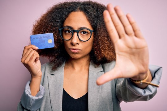 Young african american businesswoman with afro hair holding credit card over pink background with open hand doing stop sign with serious and confident expression, defense gesture