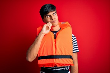 Young handsome man wearing orange safety life jacket over isolated red background with hand on chin thinking about question, pensive expression. Smiling with thoughtful face. Doubt concept.