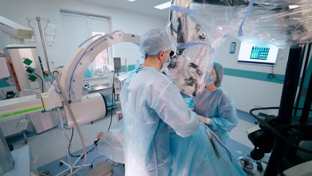 Surgeons doing an operation to a patient through the microscope. Doctors in blue protective uniform on the modern surgery cabinet background.