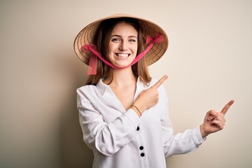 Young beautiful redhead woman wearing asian traditional conical hat over white background smiling and looking at the camera pointing with two hands and fingers to the side.