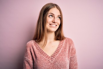 Young beautiful redhead woman wearing casual sweater over isolated pink background looking away to side with smile on face, natural expression. Laughing confident.
