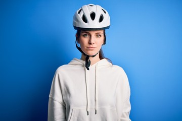 Young beautiful redhead cyclist woman wearing bike helmet over isolated blue background with serious expression on face. Simple and natural looking at the camera.