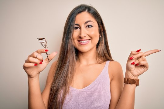 Young beautiful brunette woman holding eyelash curler over isolated white background very happy pointing with hand and finger to the side