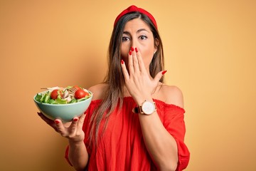 Young beautiful brunette woman eating healthy salad over isolated yellow background cover mouth...