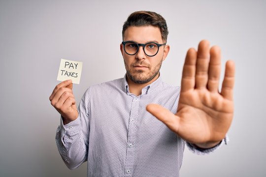 Young business man with blue eyes holding pay taxes word on paper note with open hand doing stop sign with serious and confident expression, defense gesture