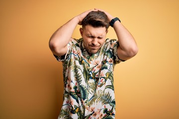 Young man with blue eyes on vacation wearing floral summer shirt over yellow background suffering from headache desperate and stressed because pain and migraine. Hands on head.