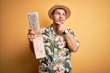 Young man on vacation wearing summer hat holding and looking at tourist map over yellow background...