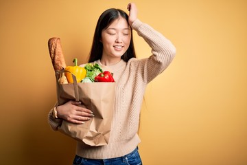 Young asian woman holding paper bag of fresh healthy groceries over yellow isolated background smiling confident touching hair with hand up gesture, posing attractive and fashionable