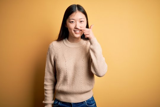 Young beautiful asian woman wearing casual sweater over yellow isolated background Pointing with hand finger to face and nose, smiling cheerful. Beauty concept