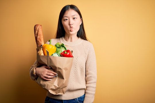 Young asian woman holding paper bag of fresh healthy groceries over yellow isolated background making fish face with lips, crazy and comical gesture. Funny expression.