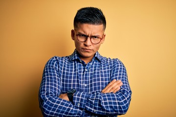 Young handsome latin man wearing casual shirt and glasses over yellow background skeptic and...