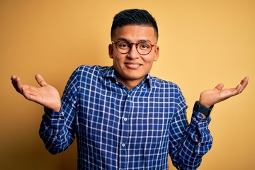 Young handsome latin man wearing casual shirt and glasses over yellow background clueless and...
