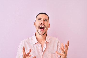 Young handsome man wearing pink shirt standing over isolated background crazy and mad shouting and yelling with aggressive expression and arms raised. Frustration concept.