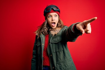 Young blonde girl wearing ski glasses and winter coat for ski weather over red background Pointing with finger surprised ahead, open mouth amazed expression, something on the front