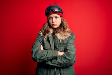 Young blonde girl wearing ski glasses and winter coat for ski weather over red background skeptic and nervous, disapproving expression on face with crossed arms. Negative person.