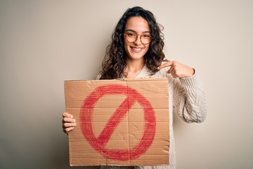Young beautiful woman with curly hair holding banner with prohibited signal message with surprise...