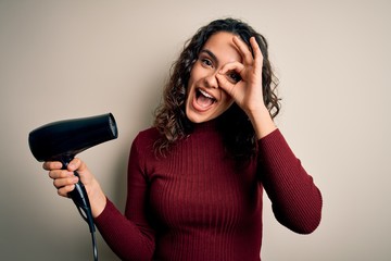 Young beautiful woman with curly hair using hair dryer over isolated white background with happy face smiling doing ok sign with hand on eye looking through fingers