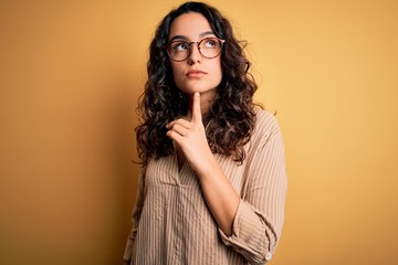 Beautiful woman with curly hair wearing striped shirt and glasses over yellow background Thinking...