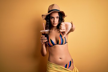 Young beautiful woman with curly hair on vacation wearing bikini drinking cola beverage with angry face, negative sign showing dislike with thumbs down, rejection concept