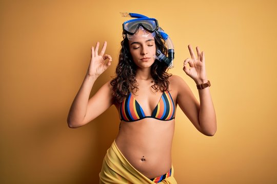 Young beautiful woman with curly hair on vacation wearing bikini and diving googles relax and smiling with eyes closed doing meditation gesture with fingers. Yoga concept.