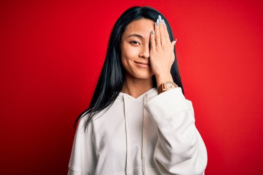 Young beautiful chinese sporty woman wearing sweatshirt over isolated red background covering one eye with hand, confident smile on face and surprise emotion.