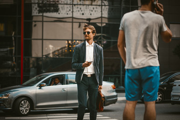 Confident young businessman in a blue sute with briefcase using cell phone or smartphone in the city