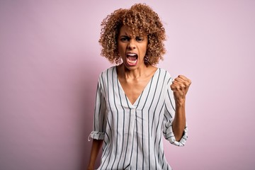 Beautiful african american woman with curly hair wearing striped t-shirt over pink background angry...